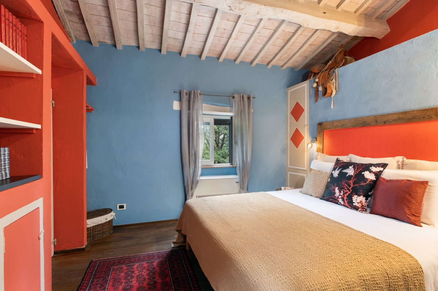 Cozy bedroom interior featuring a double bed with decorative pillows, vibrant orange and blue walls, wooden ceiling beams, and a window with natural light. The room includes a bookshelf and a rug, creating a warm and inviting atmosphere. Ideal for relaxation and comfort. | Only on SopranoVillas