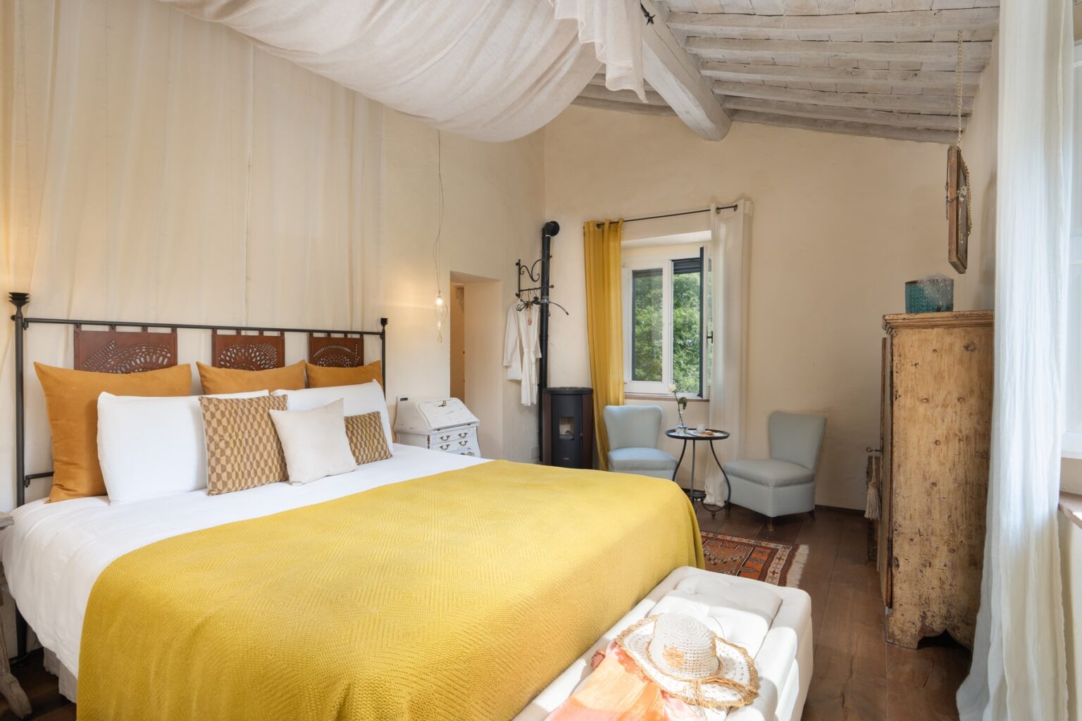 Cozy and elegantly designed bedroom featuring a king-size bed with a mustard yellow blanket, decorative pillows, and natural light streaming through a window. The room includes stylish furniture, a small seating area, and rustic wooden accents, creating a warm and inviting atmosphere. | Only on SopranoVillas