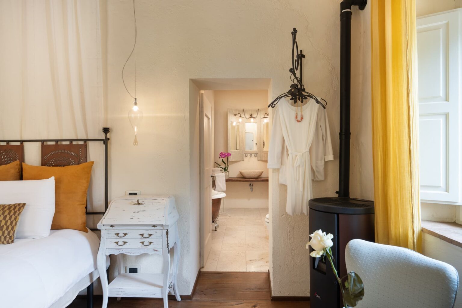 Cozy bedroom featuring a stylish iron bed with decorative pillows, a vintage white nightstand, and a soft robe hanging nearby. The room leads to a well-lit bathroom with a modern sink and a touch of greenery from a blooming orchid. Warm yellow curtains add a cheerful vibe to the serene space. | Only on SopranoVillas