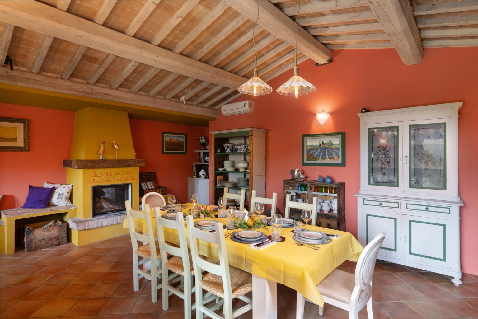Cozy dining room featuring a large wooden table set with a yellow tablecloth, surrounded by white chairs. The room has rustic wooden beams and a warm orange wall, complemented by a fireplace and a vintage cabinet. Perfect for family gatherings and meals. | Only on SopranoVillas