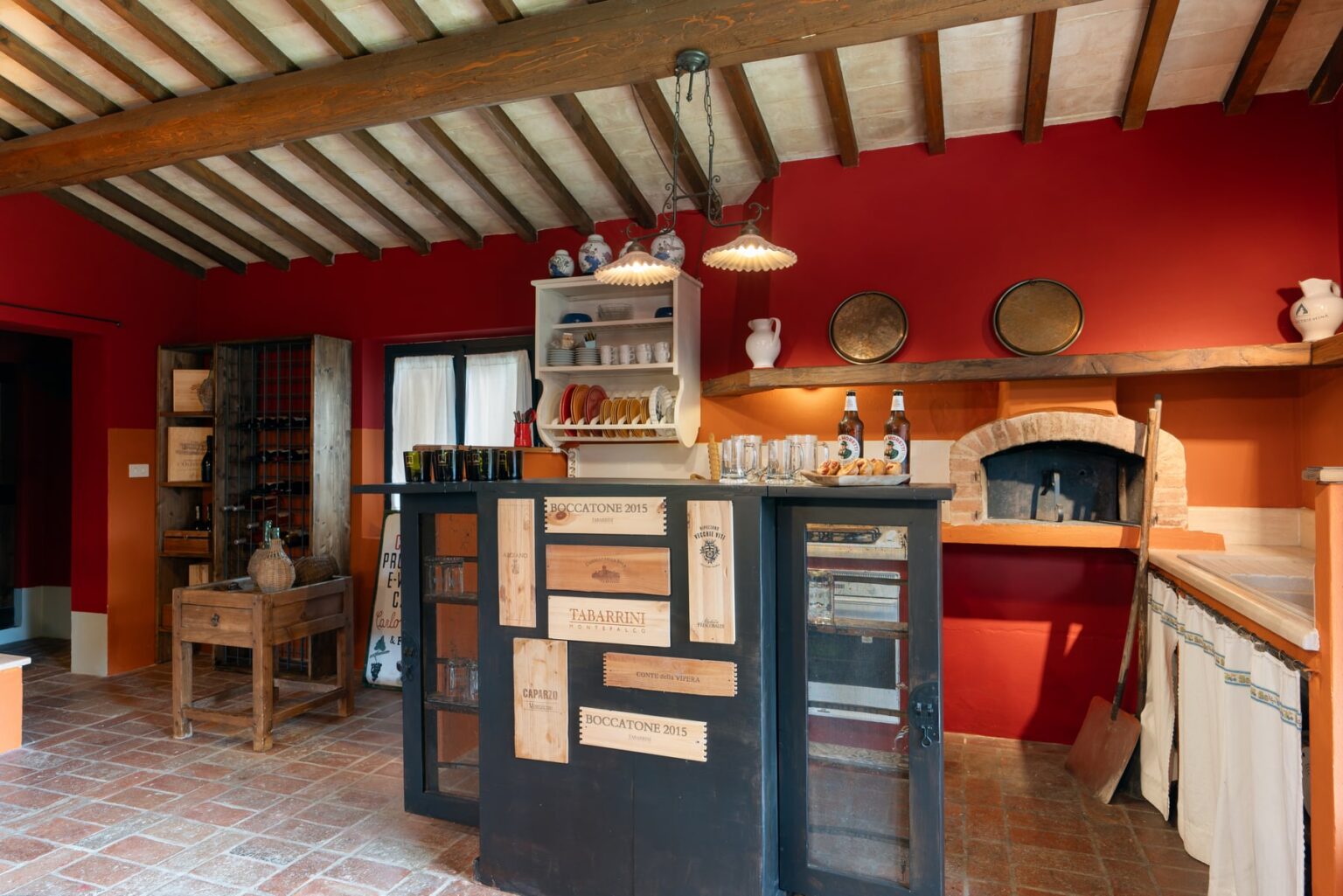 Rustic kitchen interior featuring a red accent wall, wooden beams, and a central island with glass-fronted cabinets. The space includes a traditional oven, shelves displaying pottery, and a warm, inviting atmosphere perfect for cooking and entertaining. | Only on SopranoVillas