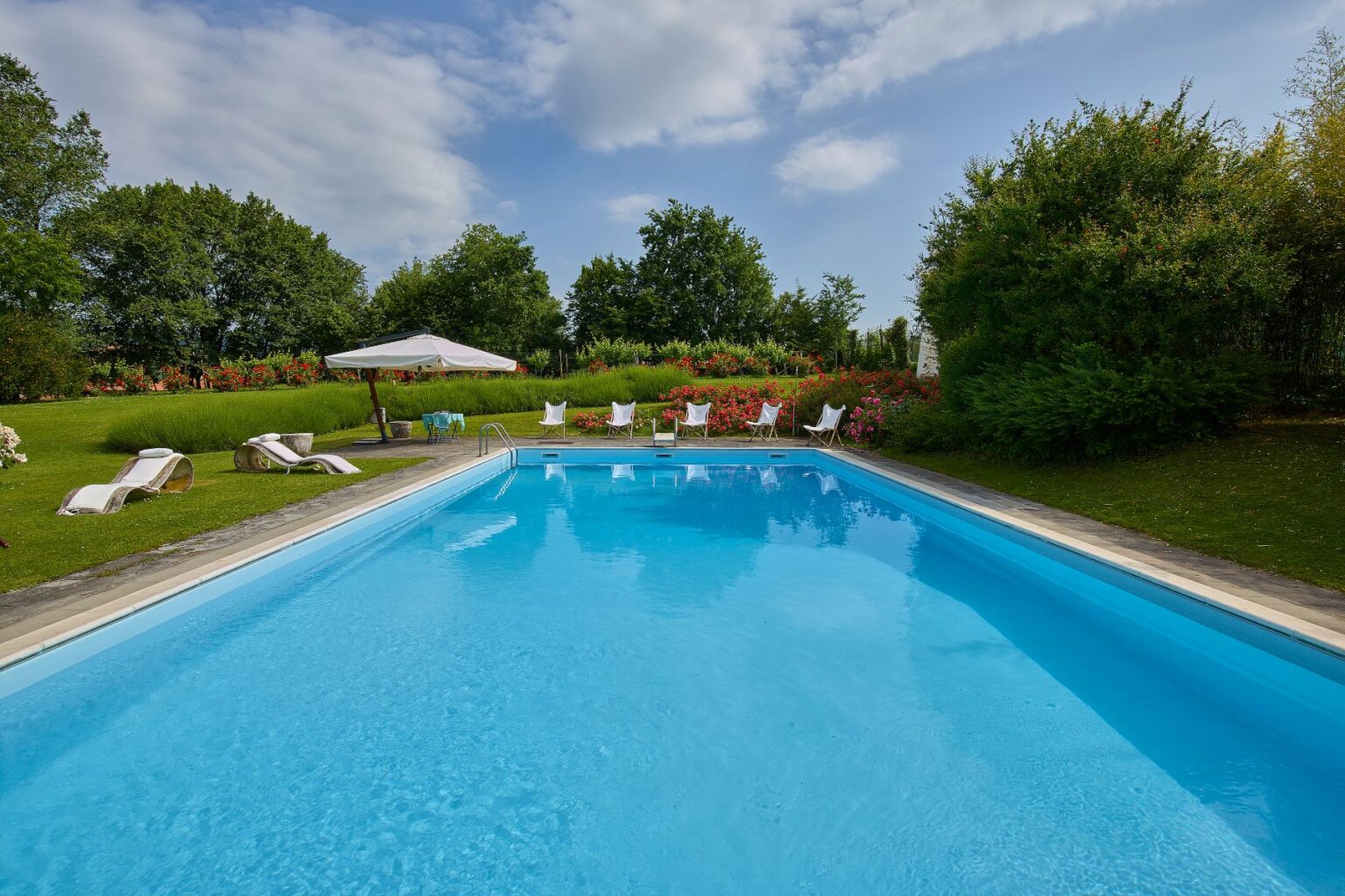 Inviting outdoor swimming pool surrounded by lush greenery and vibrant flower beds, featuring lounge chairs and a shaded umbrella for relaxation on a sunny day. | Only on SopranoVillas
