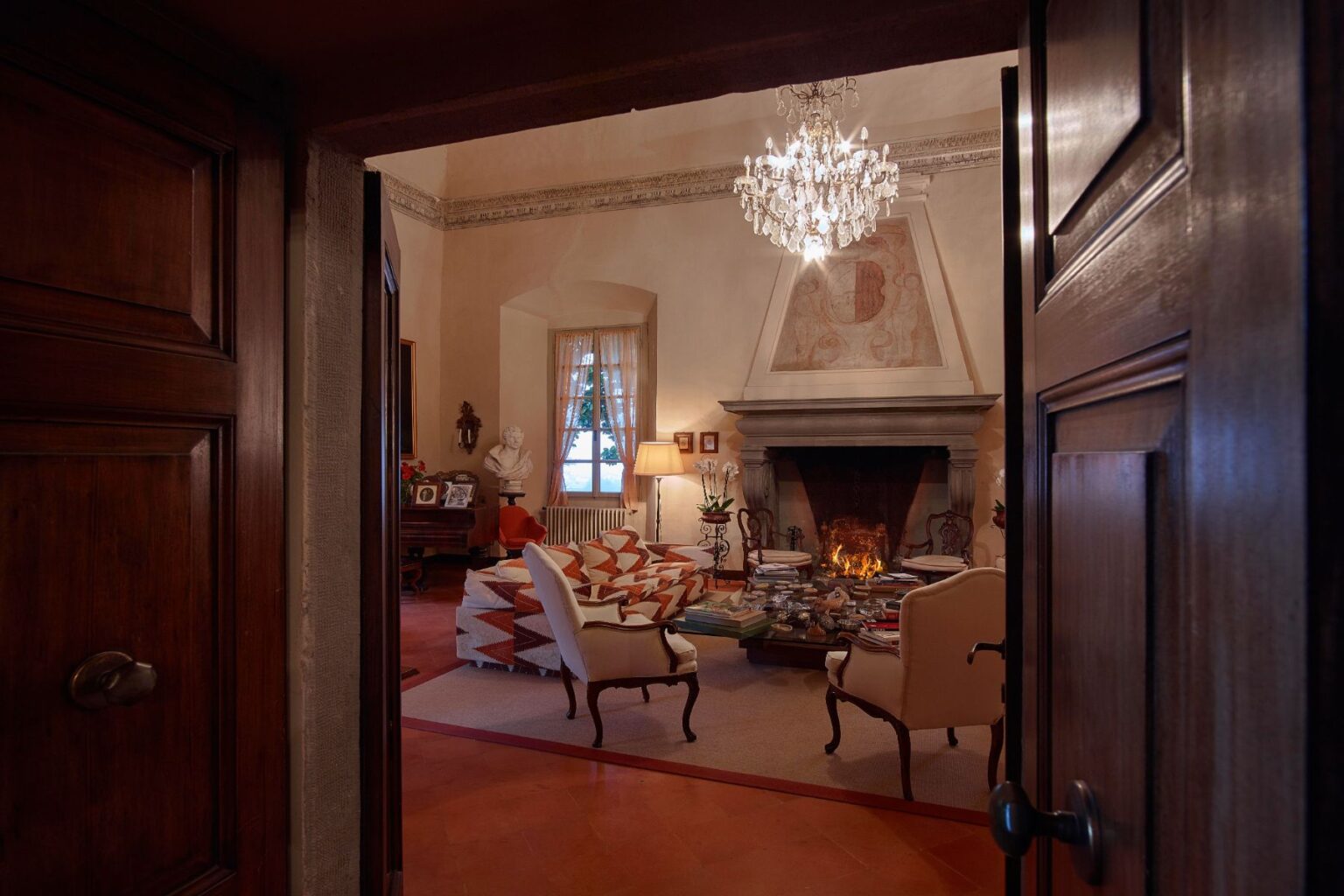 Cozy living room with a fireplace, elegant chandelier, and vintage decor, viewed from an open wooden door. The room features comfortable seating, a coffee table stacked with books, and a classic bust sculpture, creating an inviting atmosphere. | Only on SopranoVillas