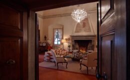 Cozy living room with a fireplace, elegant chandelier, and vintage decor, viewed from an open wooden door. The room features comfortable seating, a coffee table stacked with books, and a classic bust sculpture, creating an inviting atmosphere.