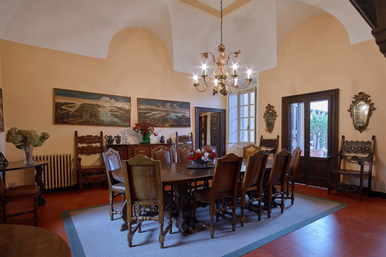 Elegant dining room featuring a long wooden table surrounded by ornate chairs, complemented by a chandelier and vintage artwork on the walls. Natural light streams in through large windows, enhancing the warm, inviting atmosphere. | Only on SopranoVillas
