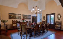 Elegant dining room featuring a long wooden table surrounded by ornate chairs, complemented by a chandelier and vintage artwork on the walls. Natural light streams in through large windows, enhancing the warm, inviting atmosphere.
