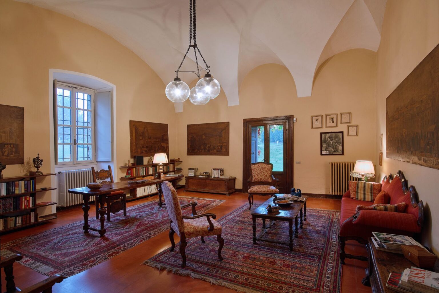 Cozy interior of a vintage living room featuring elegant furniture, a wooden table, and decorative paintings on the walls. The room is illuminated by a modern chandelier and has a warm color palette, creating an inviting atmosphere. A large window allows natural light to fill the space, enhancing the traditional decor. | Only on SopranoVillas