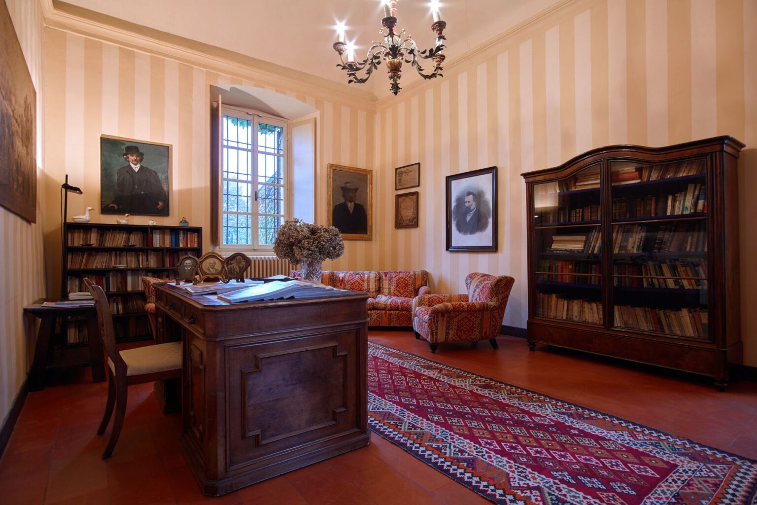 Cozy study room featuring a wooden desk, vintage armchairs, and a large bookshelf filled with books. The room is adorned with framed portraits and has a window that lets in natural light, along with a decorative chandelier and a patterned rug on the floor. | Only on SopranoVillas