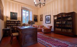 Cozy study room featuring a wooden desk, vintage armchairs, and a large bookshelf filled with books. The room is adorned with framed portraits and has a window that lets in natural light, along with a decorative chandelier and a patterned rug on the floor.