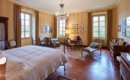 Spacious and elegantly decorated bedroom featuring a large bed, vintage furniture, and ample natural light from tall windows. The room includes a cozy seating area, a writing desk, and decorative artwork on the walls, creating a warm and inviting atmosphere.