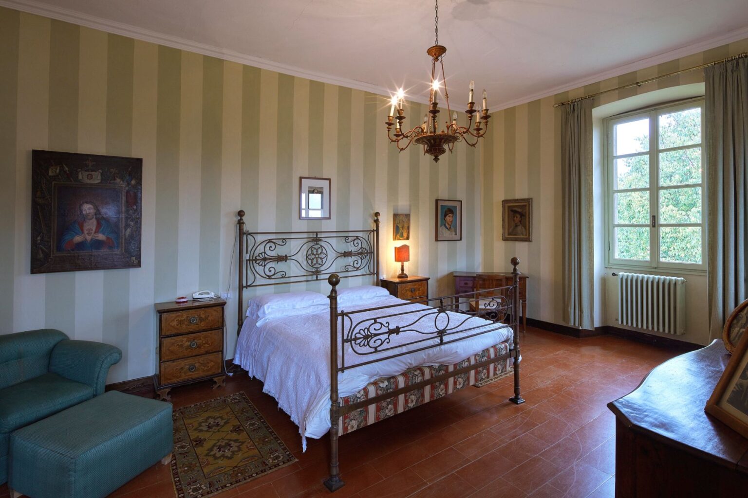 Vintage bedroom featuring a wrought iron bed, antique furniture, and striped walls. The room includes a cozy seating area, a chandelier, and framed artwork, with natural light pouring in from a large window. Ideal for a tranquil stay or a romantic getaway. | Only on SopranoVillas