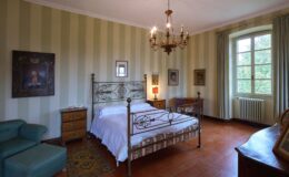 Vintage bedroom featuring a wrought iron bed, antique furniture, and striped walls. The room includes a cozy seating area, a chandelier, and framed artwork, with natural light pouring in from a large window. Ideal for a tranquil stay or a romantic getaway.