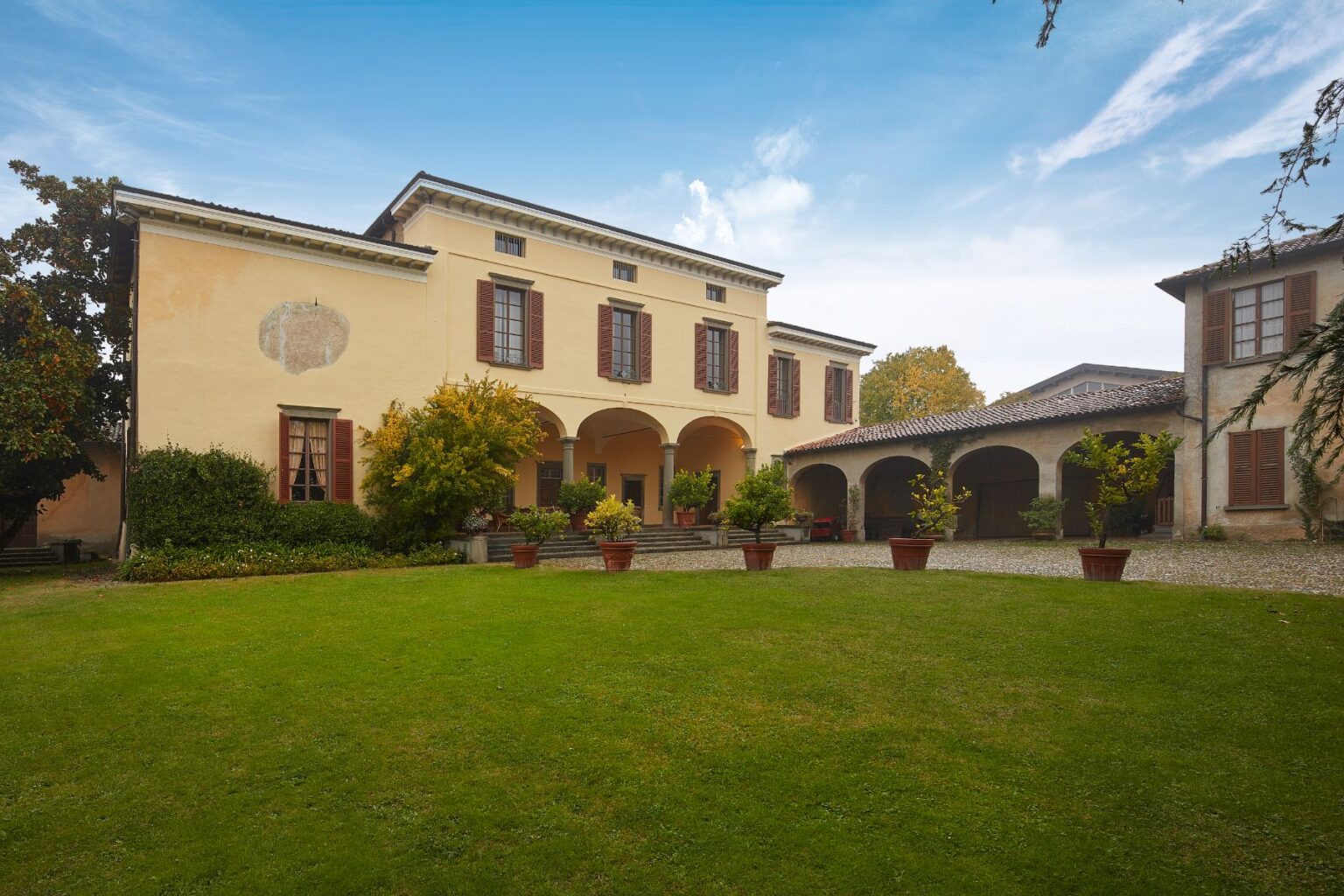 Historic villa with a spacious garden, featuring arched entryways and potted plants, set against a clear blue sky. The architecture showcases classic design elements with wooden shutters and a charming courtyard. | Only on SopranoVillas