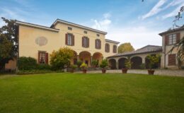 Historic villa with a spacious garden, featuring arched entryways and potted plants, set against a clear blue sky. The architecture showcases classic design elements with wooden shutters and a charming courtyard.