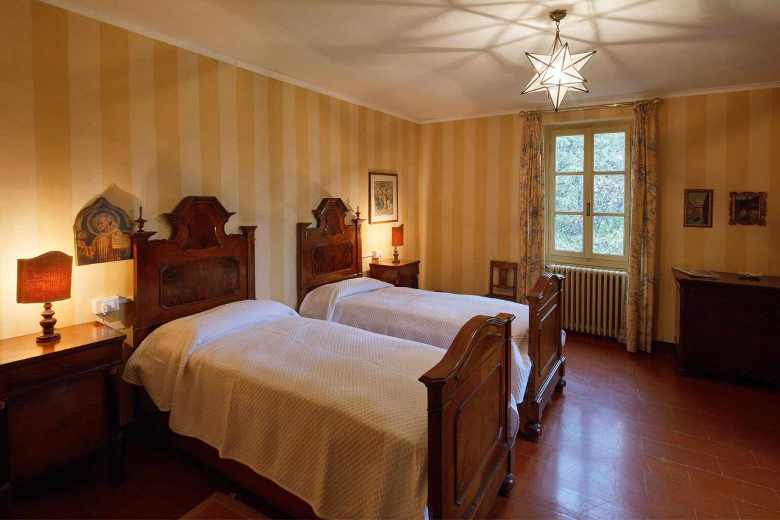 Cozy guest room featuring two antique wooden beds with white bedding, a bedside lamp, and striped yellow walls. A window offers natural light, while decorative art and a wooden cabinet enhance the inviting atmosphere. Ideal for relaxing stays. | Only on SopranoVillas