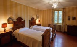 Cozy guest room featuring two antique wooden beds with white bedding, a bedside lamp, and striped yellow walls. A window offers natural light, while decorative art and a wooden cabinet enhance the inviting atmosphere. Ideal for relaxing stays.