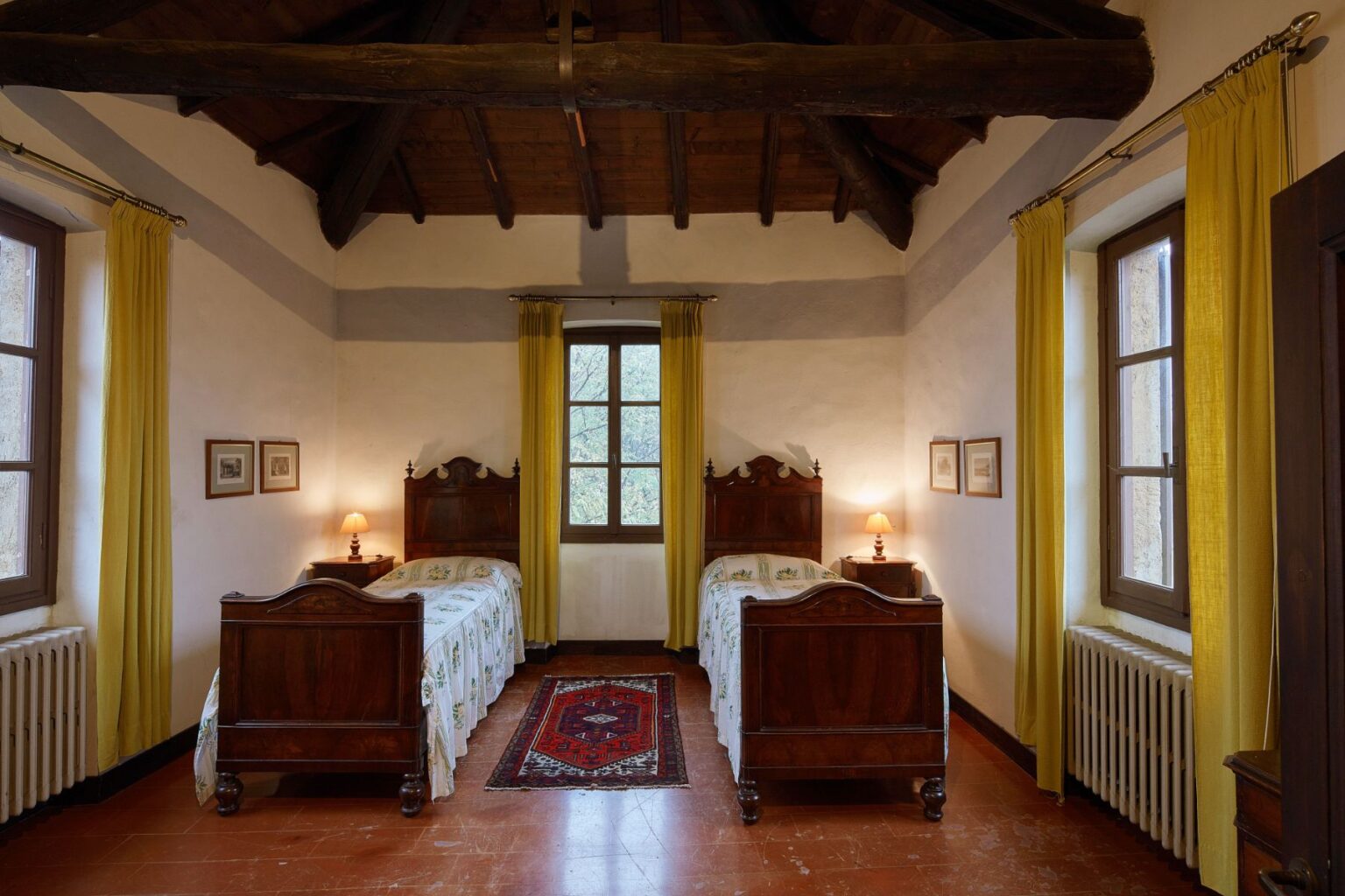 Cozy guest room featuring two wooden twin beds with floral bedding, yellow curtains, and rustic beams. The room is illuminated by a bedside lamp and showcases framed artwork on the walls, offering a warm and inviting atmosphere. | Only on SopranoVillas
