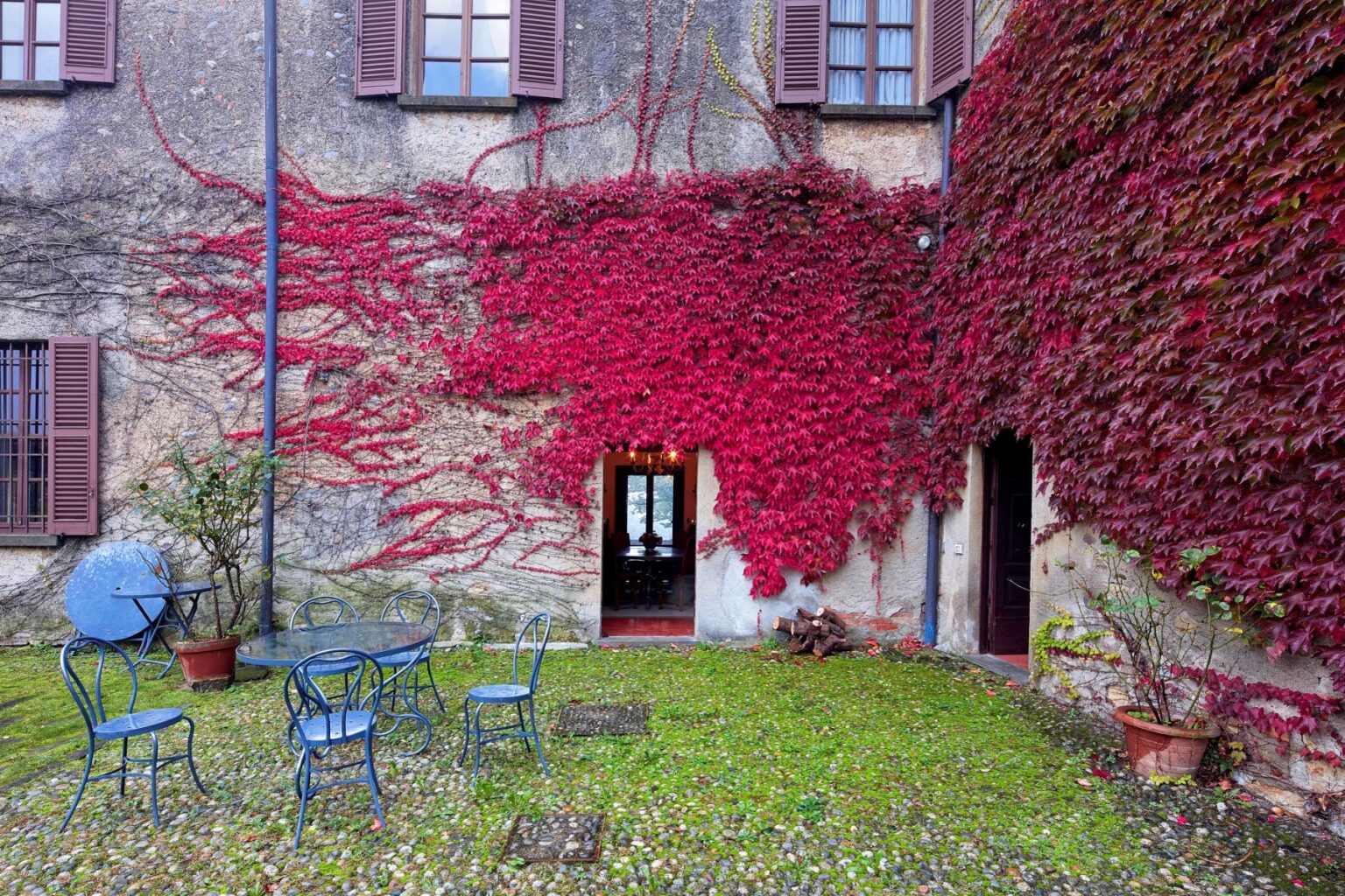 Charming courtyard featuring a rustic stone wall adorned with vibrant red ivy, two entrances, and a small seating area with blue metal chairs and tables surrounded by lush greenery. | Only on SopranoVillas