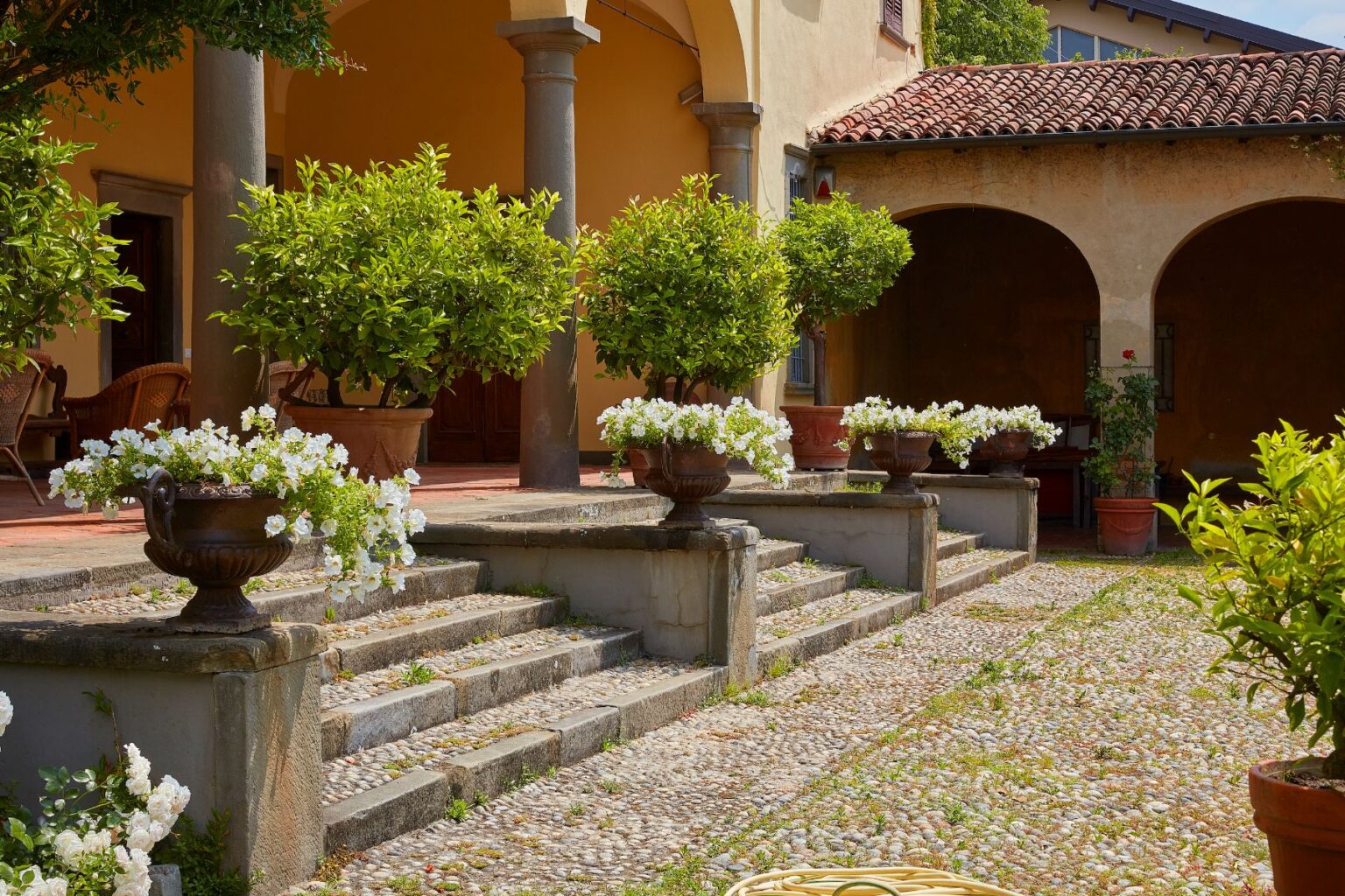 Beautiful courtyard with stone steps leading to a rustic building, adorned with lush green potted plants and vibrant white flowers in decorative urns, creating a serene and inviting atmosphere. | Only on SopranoVillas