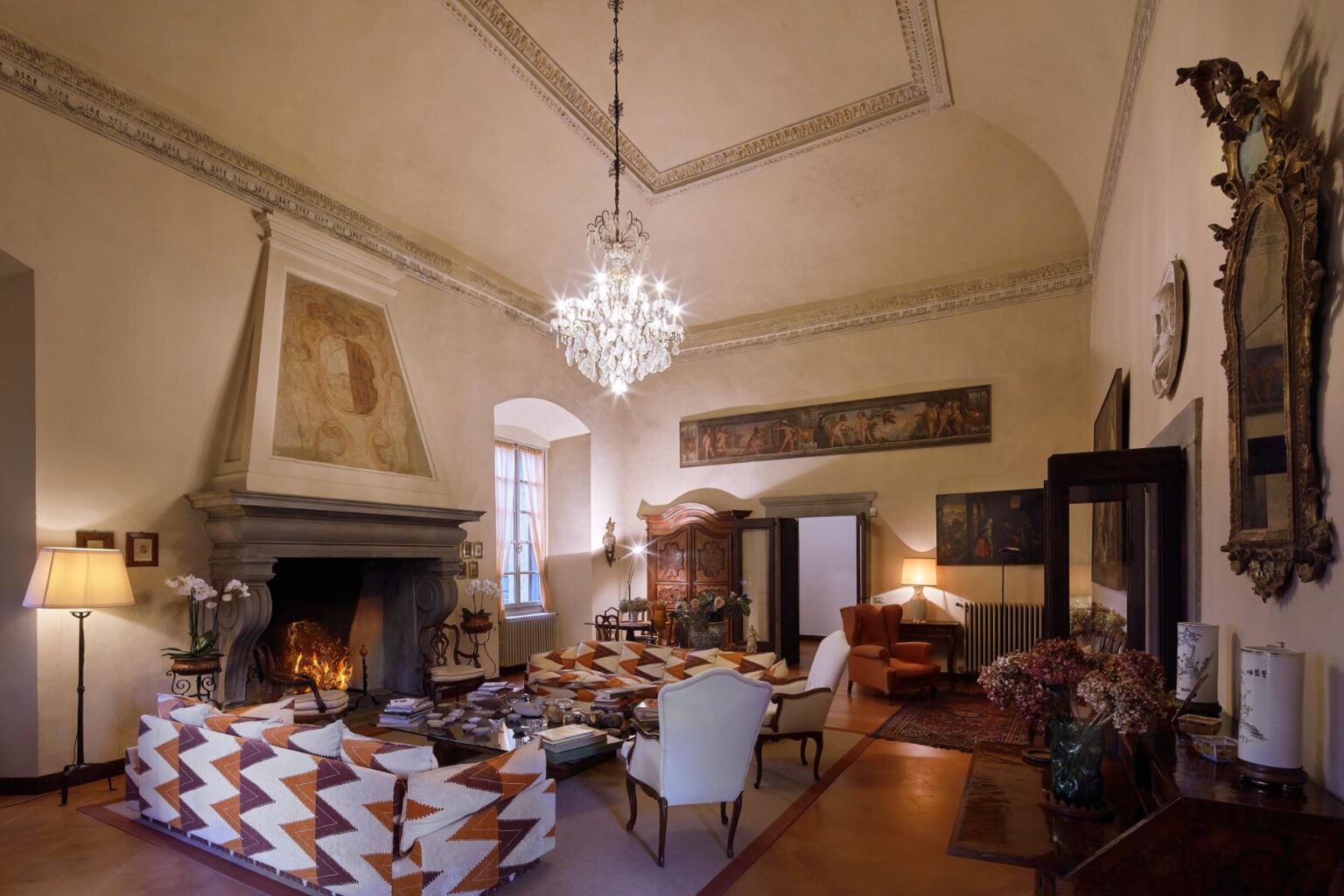 Elegant living room featuring a high ceiling with intricate moldings, a crystal chandelier, and a cozy fireplace. The space is adorned with vintage furniture, including patterned sofas and a classic wooden cabinet. Natural light filters through large windows, enhancing the warm ambiance. Decorative paintings and floral arrangements complete the sophisticated decor. | Only on SopranoVillas
