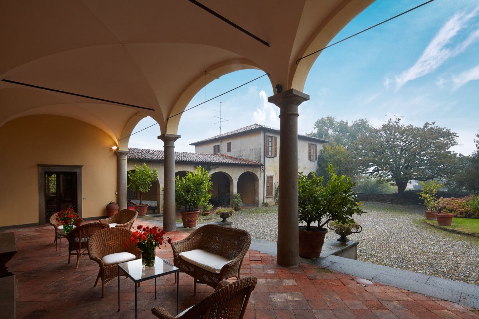 Cozy outdoor patio with wicker chairs and vibrant flower arrangements, overlooking a rustic Italian villa surrounded by lush greenery and a gravel courtyard. Ideal for relaxation in a serene countryside setting. | Only on SopranoVillas