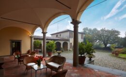 Cozy outdoor patio with wicker chairs and vibrant flower arrangements, overlooking a rustic Italian villa surrounded by lush greenery and a gravel courtyard. Ideal for relaxation in a serene countryside setting.