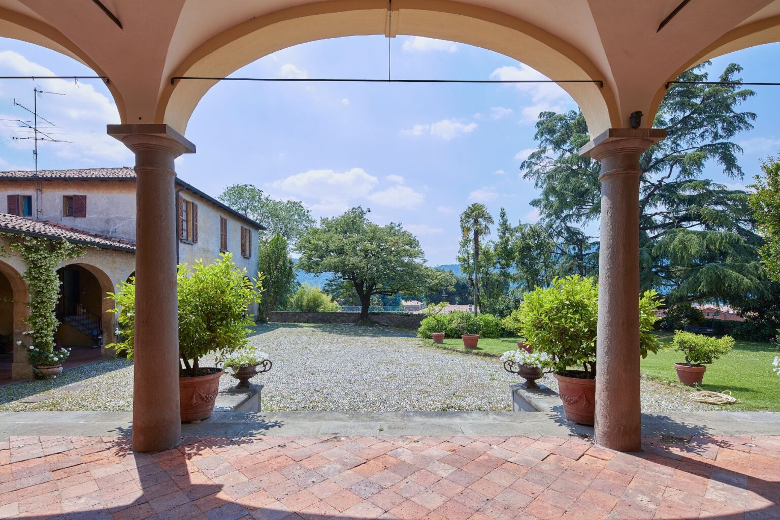 View from an arched porch showcasing a serene garden with potted plants and a gravel pathway, leading to a rustic Italian villa surrounded by trees under a clear blue sky. Perfect for travelers seeking a picturesque outdoor setting. | Only on SopranoVillas
