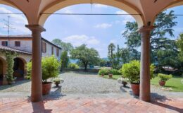 View from an arched porch showcasing a serene garden with potted plants and a gravel pathway, leading to a rustic Italian villa surrounded by trees under a clear blue sky. Perfect for travelers seeking a picturesque outdoor setting.