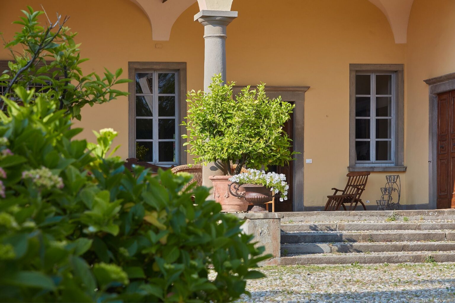 Charming entrance of a villa featuring a yellow exterior, decorative potted plants, and stone steps leading to wooden doors, surrounded by lush greenery and sunlight. | Only on SopranoVillas