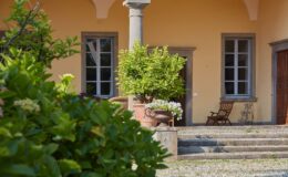 Charming entrance of a villa featuring a yellow exterior, decorative potted plants, and stone steps leading to wooden doors, surrounded by lush greenery and sunlight.