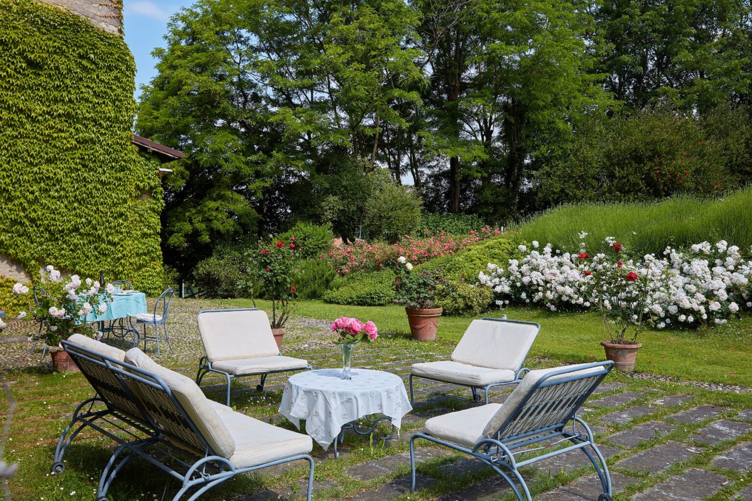 Outdoor garden seating area featuring four lounge chairs and a small table with a vase of pink flowers, surrounded by lush greenery and blooming rose bushes, creating a serene and inviting atmosphere. | Only on SopranoVillas