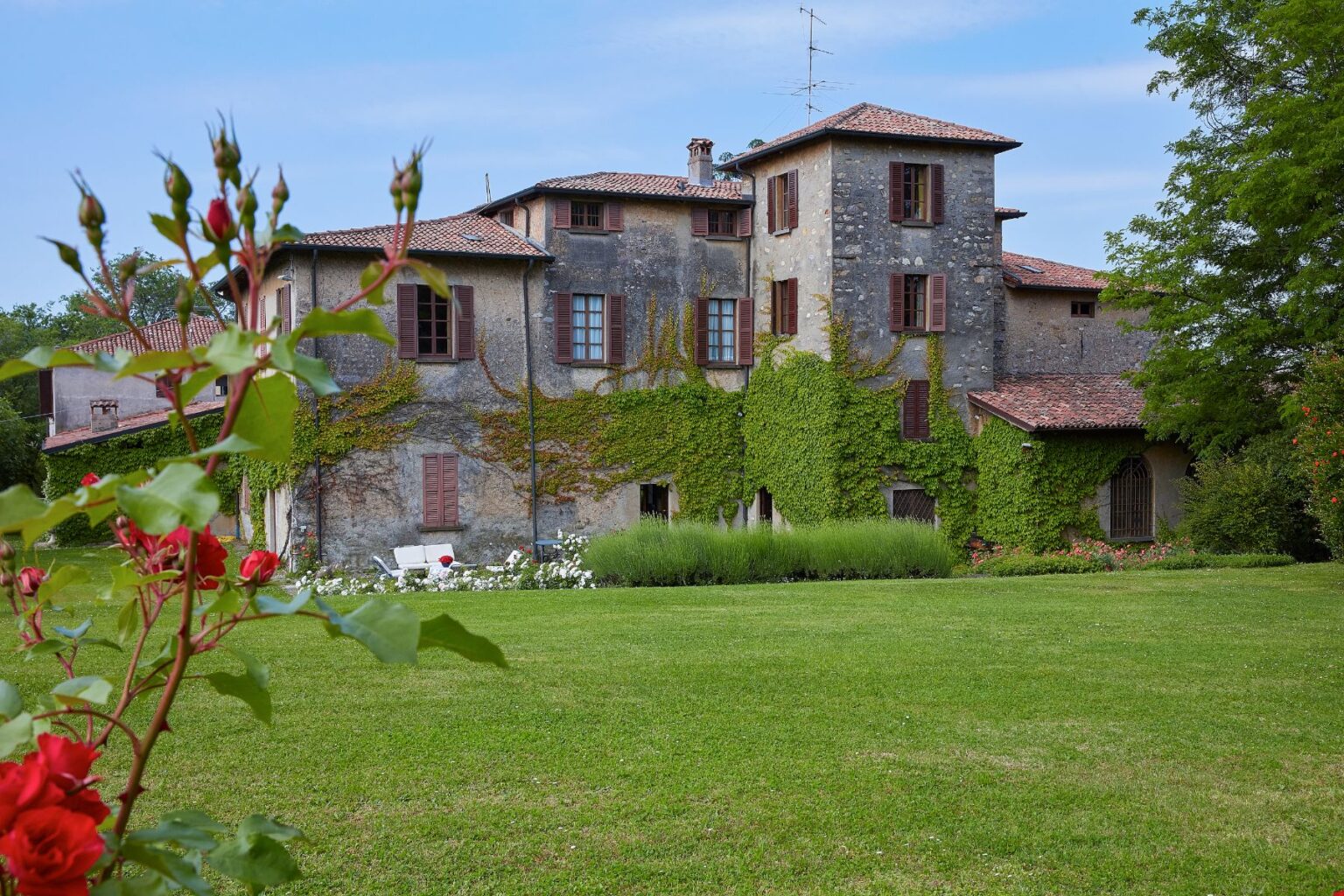 Historic villa surrounded by lush gardens and blooming roses, featuring ivy-covered walls and traditional architecture under a clear blue sky. Ideal for nature lovers and photography enthusiasts. | Only on SopranoVillas