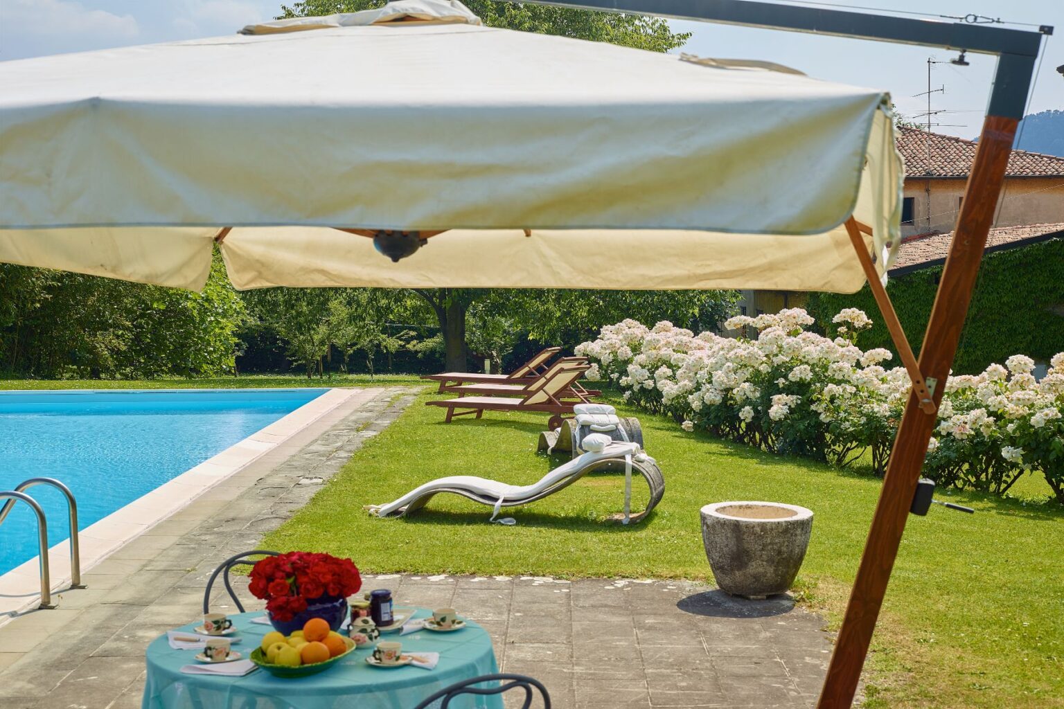A serene outdoor scene featuring a swimming pool surrounded by lush greenery and blooming white hydrangeas. In the foreground, a table set with tea cups and fresh fruits sits under a large umbrella, while lounge chairs are positioned nearby for relaxation. The setting exudes tranquility, perfect for a summer getaway. | Only on SopranoVillas