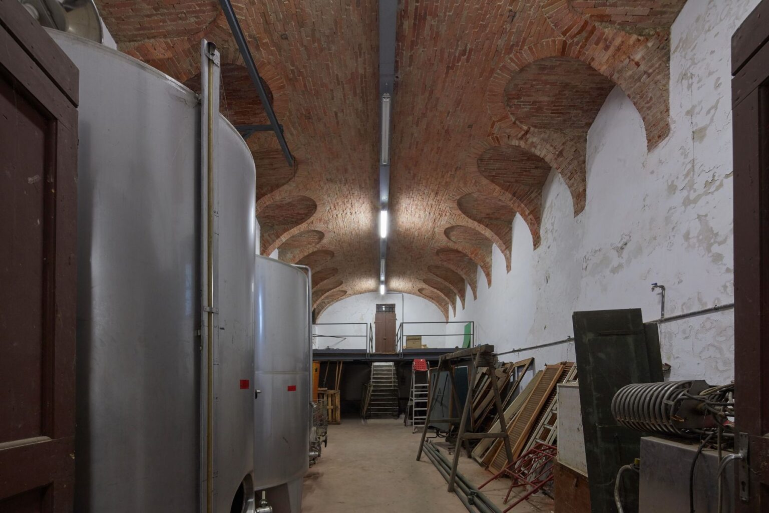 Interior view of a spacious warehouse featuring a high brick arched ceiling, metal storage tanks on the left, and various wooden and metal items stacked against the right wall, with a staircase leading to a loft area in the background. | Only on SopranoVillas