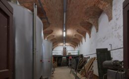Interior view of a spacious warehouse featuring a high brick arched ceiling, metal storage tanks on the left, and various wooden and metal items stacked against the right wall, with a staircase leading to a loft area in the background.