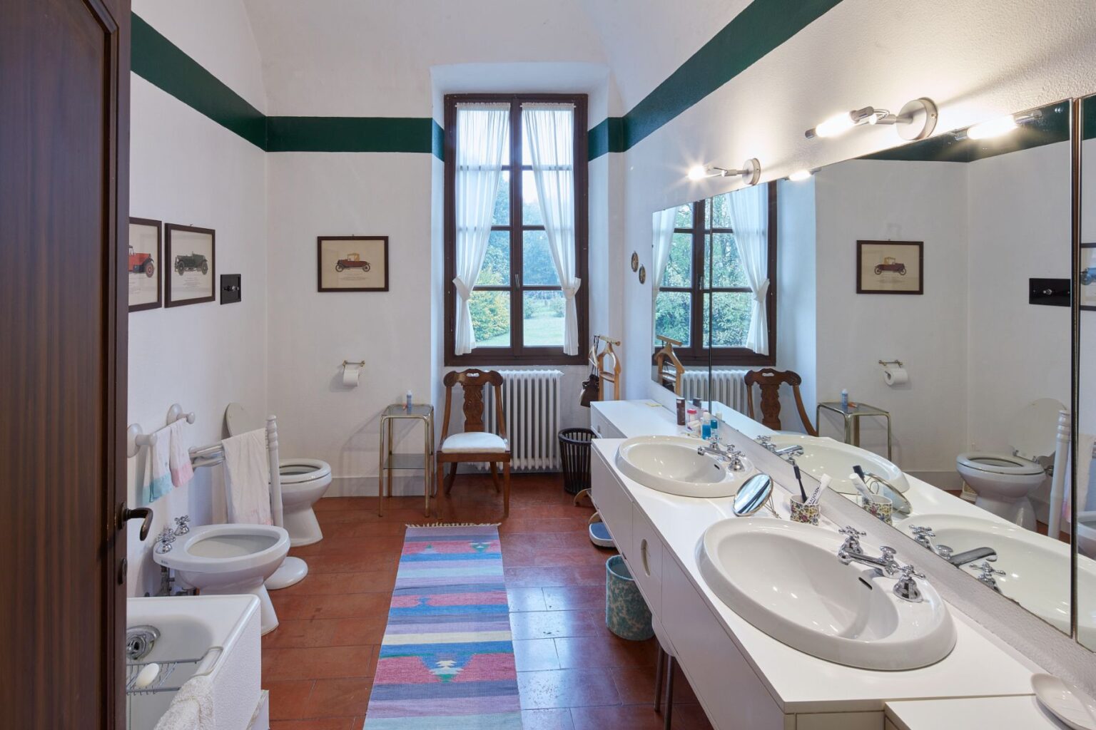 Bright and spacious bathroom featuring dual sinks, a large mirror, and natural light from windows. Decorated with vintage car prints, a colorful rug, and a wooden chair, the room includes a toilet and bidet, emphasizing a blend of modern and classic design elements. | Only on SopranoVillas