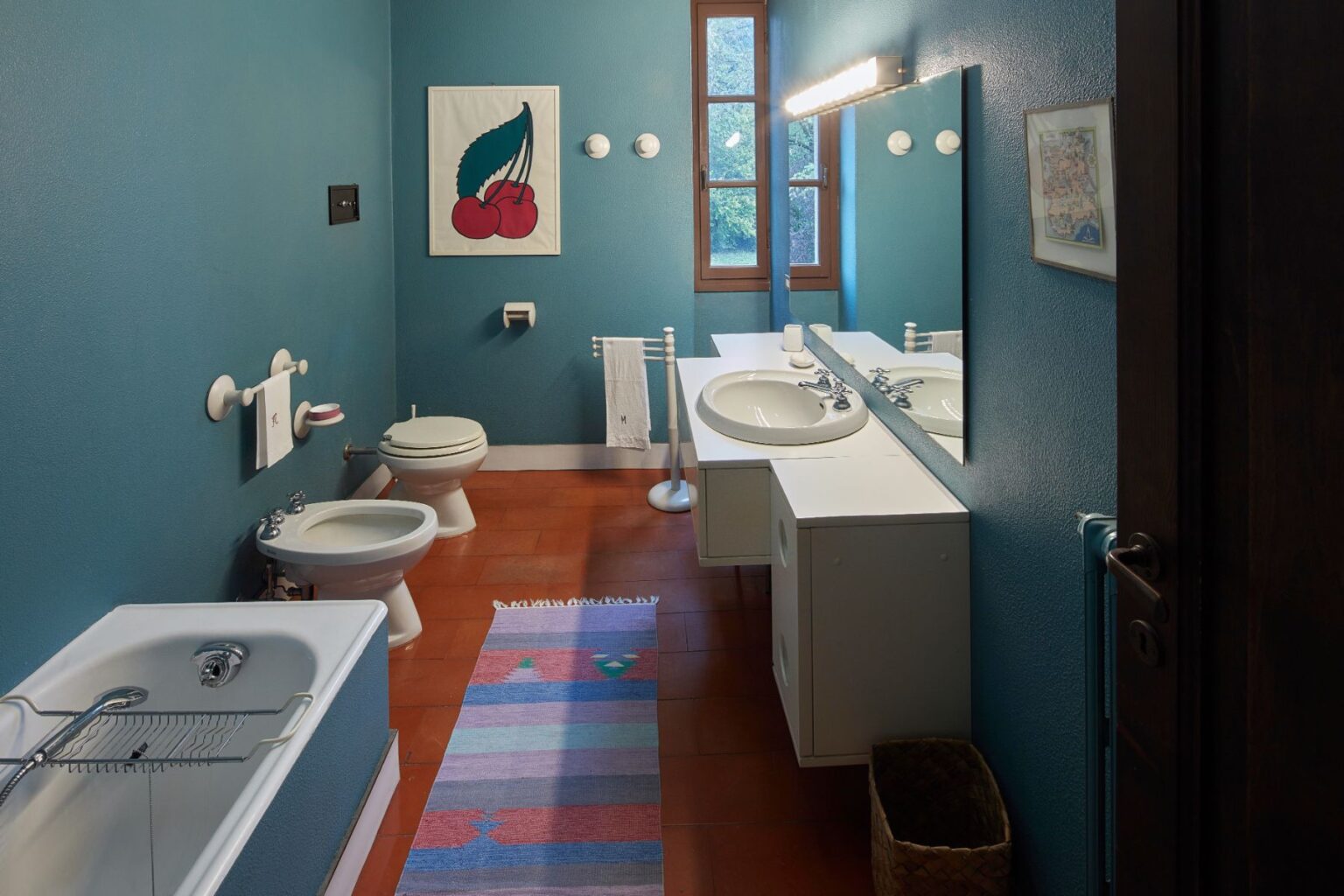 Modern bathroom with teal walls, a bathtub, bidet, and toilet. Features a double sink vanity with a large mirror, decorative cherry artwork, and a colorful rug on the terracotta floor. Natural light enters through a window, enhancing the space's inviting atmosphere. | Only on SopranoVillas