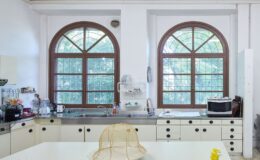 Bright and airy kitchen featuring large arched windows, stainless steel sink, and white cabinetry. The countertop displays a mesh food cover and a decorative plate, while kitchen appliances and utensils are neatly arranged. Greenery is visible outside the windows, enhancing the natural light in the space.