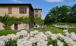 Historic villa surrounded by blooming white and pink roses, featuring a lush green garden, ivy-covered walls, and outdoor seating areas on a sunny day.