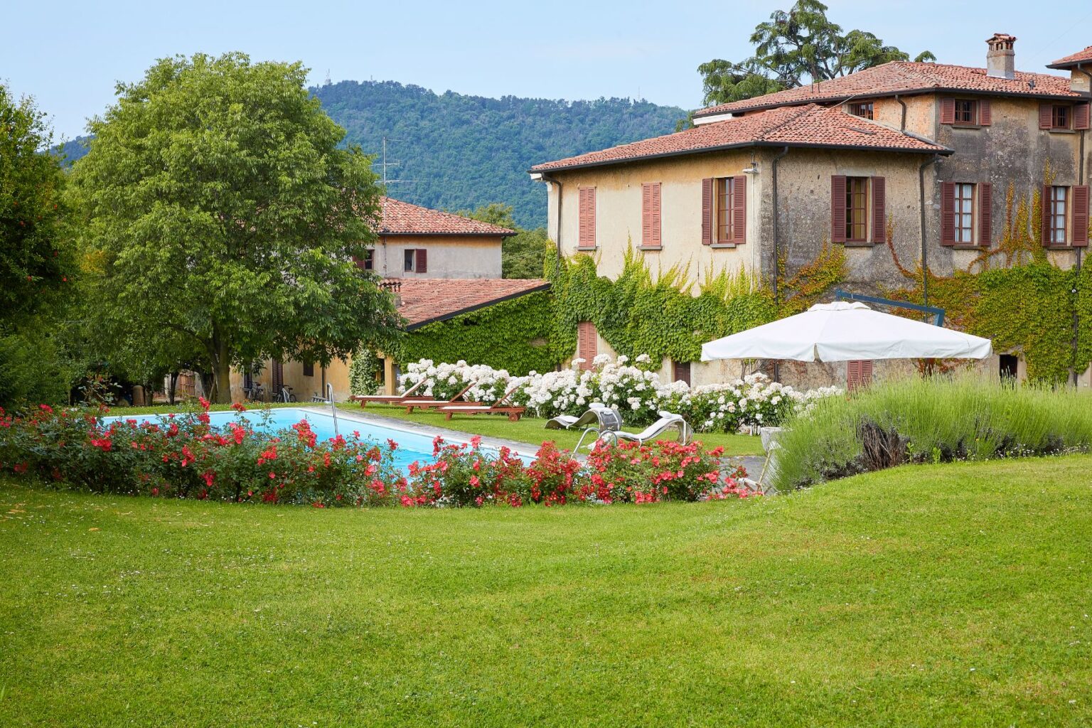 Scenic view of a serene villa with a swimming pool surrounded by lush greenery and blooming flowers, set against a backdrop of rolling hills. The image features a well-maintained garden, sun loungers, and a shaded area with an umbrella, ideal for relaxation. | Only on SopranoVillas