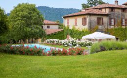 Scenic view of a serene villa with a swimming pool surrounded by lush greenery and blooming flowers, set against a backdrop of rolling hills. The image features a well-maintained garden, sun loungers, and a shaded area with an umbrella, ideal for relaxation.