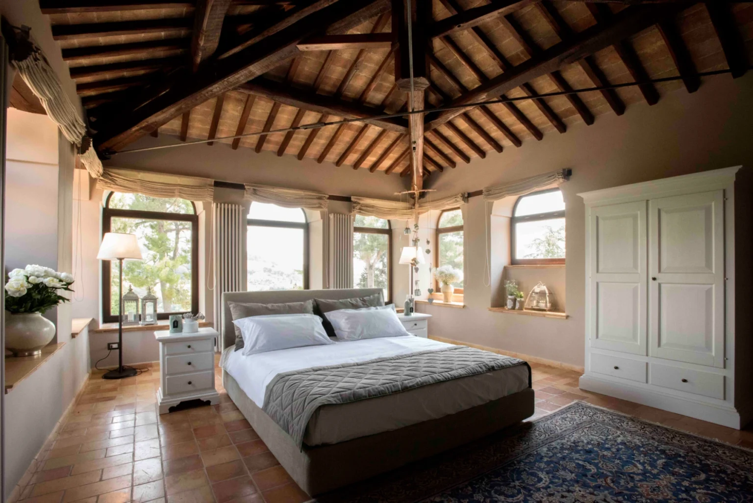 Spacious and elegantly designed bedroom featuring a king-size bed with plush bedding, wooden beams on the ceiling, large windows allowing natural light, and stylish decor elements including a bedside table and a white wardrobe. | Only on SopranoVillas