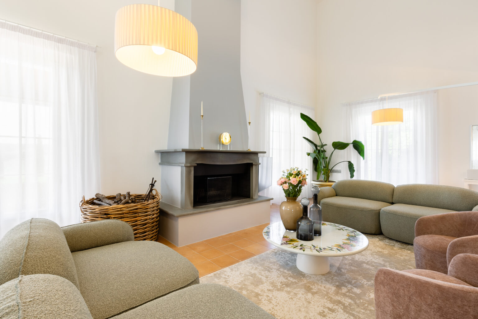 Modern living room featuring a cozy design with a gray stone fireplace, plush green and brown sofas, and a decorative round coffee table. Natural light floods the space through sheer white curtains, complemented by indoor plants and a floral centerpiece. Ideal for creating a welcoming and stylish atmosphere. | Only on SopranoVillas