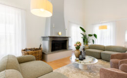 Modern living room featuring a cozy design with a gray stone fireplace, plush green and brown sofas, and a decorative round coffee table. Natural light floods the space through sheer white curtains, complemented by indoor plants and a floral centerpiece. Ideal for creating a welcoming and stylish atmosphere.