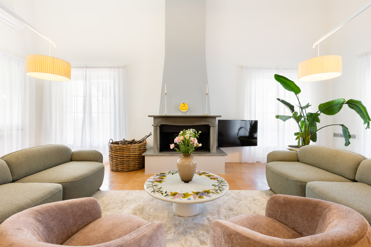 Modern living room featuring a minimalist design with a gray stone fireplace, stylish light fixtures, and comfortable seating. A round coffee table adorned with floral decor sits in the center, surrounded by soft armchairs and a large window with sheer curtains. A decorative plant adds a touch of greenery to the serene space. | Only on SopranoVillas
