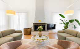 Modern living room featuring a minimalist design with a gray stone fireplace, stylish light fixtures, and comfortable seating. A round coffee table adorned with floral decor sits in the center, surrounded by soft armchairs and a large window with sheer curtains. A decorative plant adds a touch of greenery to the serene space.