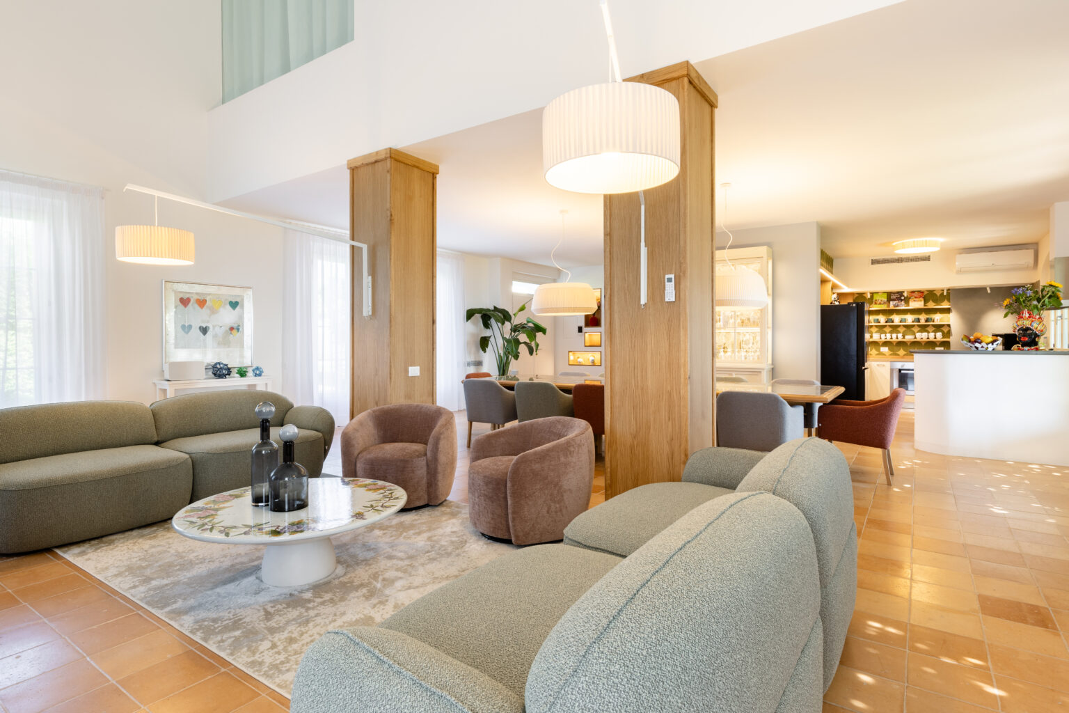 Modern living room featuring stylish furniture, including a green sofa and cozy brown armchairs, complemented by a round coffee table. Natural light floods the space through sheer curtains, creating a warm and inviting atmosphere. A contemporary kitchen is visible in the background, showcasing a sleek design and functional layout. | Only on SopranoVillas