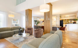 Modern living room featuring stylish furniture, including a green sofa and cozy brown armchairs, complemented by a round coffee table. Natural light floods the space through sheer curtains, creating a warm and inviting atmosphere. A contemporary kitchen is visible in the background, showcasing a sleek design and functional layout.