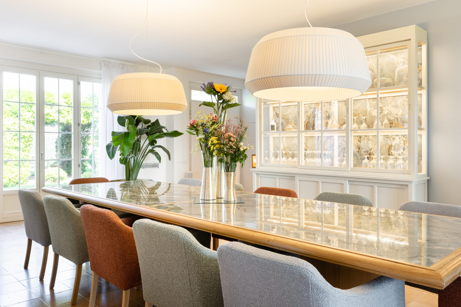Modern dining room featuring a long wooden table with a marble top, surrounded by colorful upholstered chairs. A stylish floral arrangement sits in the center, complemented by two large pendant lights overhead. A glass display cabinet filled with elegant dishware is visible in the background, along with large windows allowing natural light to illuminate the space. | Only on SopranoVillas