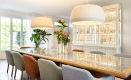 Modern dining room featuring a long wooden table with a marble top, surrounded by colorful upholstered chairs. A stylish floral arrangement sits in the center, complemented by two large pendant lights overhead. A glass display cabinet filled with elegant dishware is visible in the background, along with large windows allowing natural light to illuminate the space.